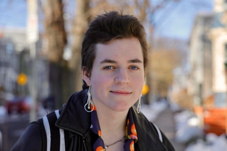 A person with short brown hair, wearing silver earrings, stands on a snowy sidewalk.