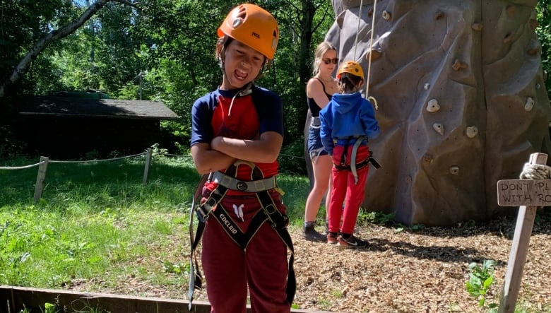 A boy stands dressed in rock climbing gear in front of a rock climbing wall. 