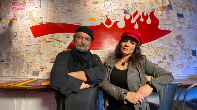 A man and a woman stand inside their restaurant in front of a wall with messages written on sticky notes by former customers.