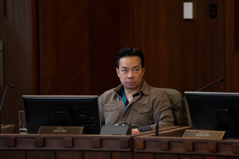 An East Asian man wearing a brown long-sleeved shirt sits in a city council's chambers.