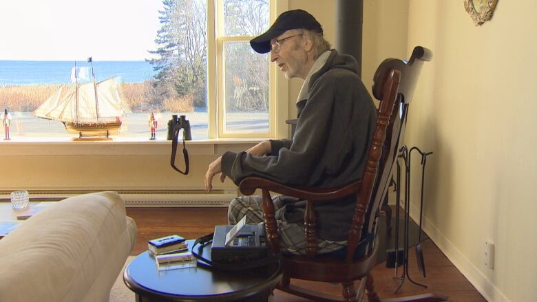 A man sits in a rocking chair by a fire and a window opening to the blue ocean. 