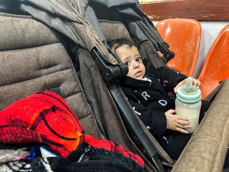 A young boy with a skin infection holds a bottle and looks out from a carseat.