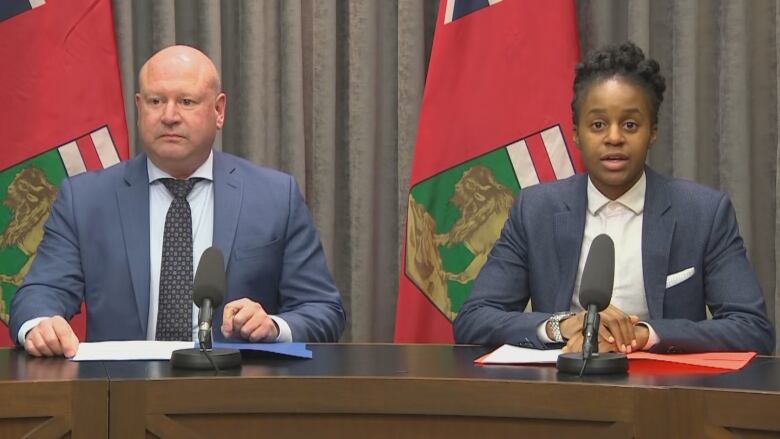 Two people sit in front of microphones with Manitoba flags behind them.