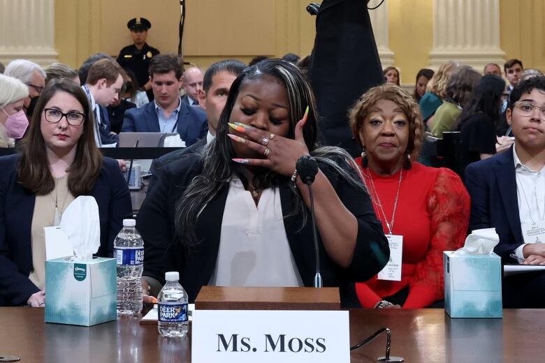 A woman with long and colorful fingernails holds a hand to the face while sitting at a table at an indoor meeting with several onlookers shown in the background.