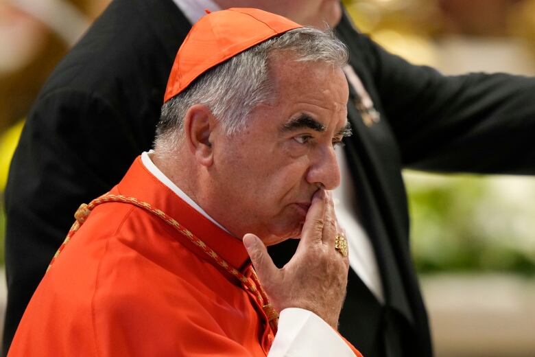 A clean-shaven older man wearing a red religious hat is shown in closeup, with his right hand resting over his mouth.