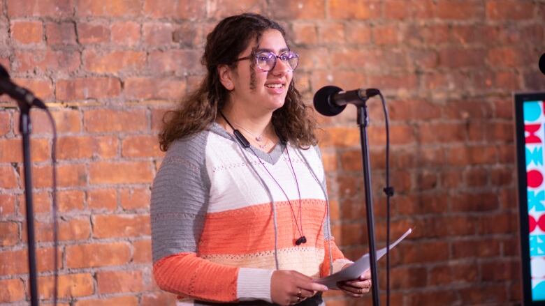 A young person in glasses holds a sheet of paper and reads into a microphone. They stand in front of a brick background.