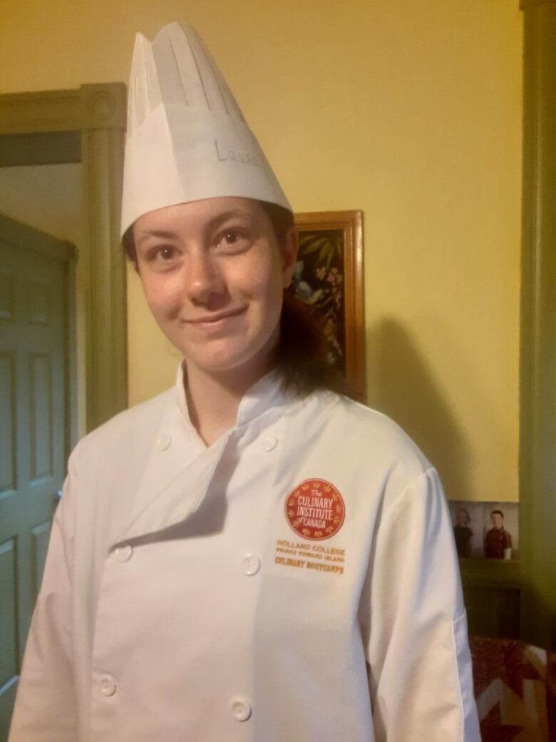 A young woman in a chef's coat and hat stands in a kitchen.