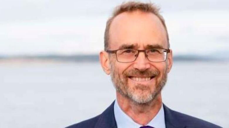 David Hilderman, a smiling white man wearing glasses and a purple tie, smiles in front of an ocean backdrop.