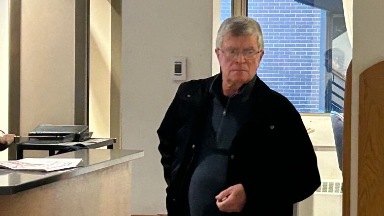 Man with short silver hair and glasses wearing black jacket over navy polo shirt. 