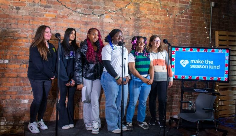 One woman and five teens stand on a stage in front of microphones.