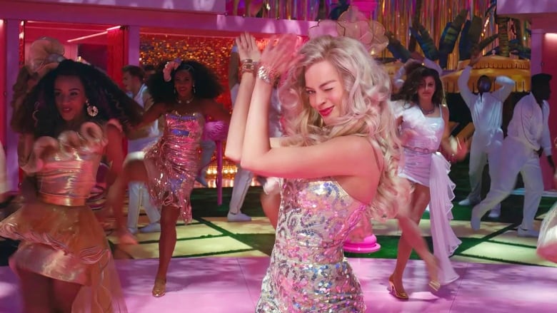 Scene from the Barbie movie. Performers dressed in sparkly party clothes dance in unison inside a pink plastic dollhouse set. Barbie, at centre, winks at the camera.