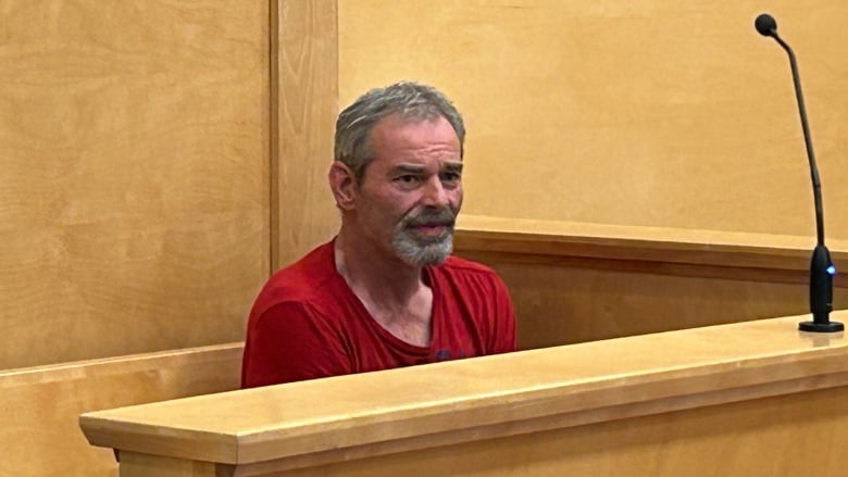 Man with grey hair and a grey goatee in a baggy red t-shirt.