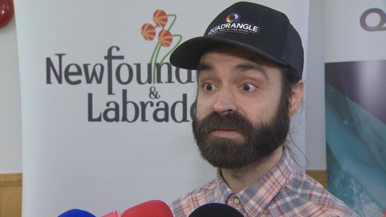 Charlie Murphy wearing a black baseball cap with Quadrangle's logo, speaking to reporters in front of a Newfoundland and Labrador banner. 