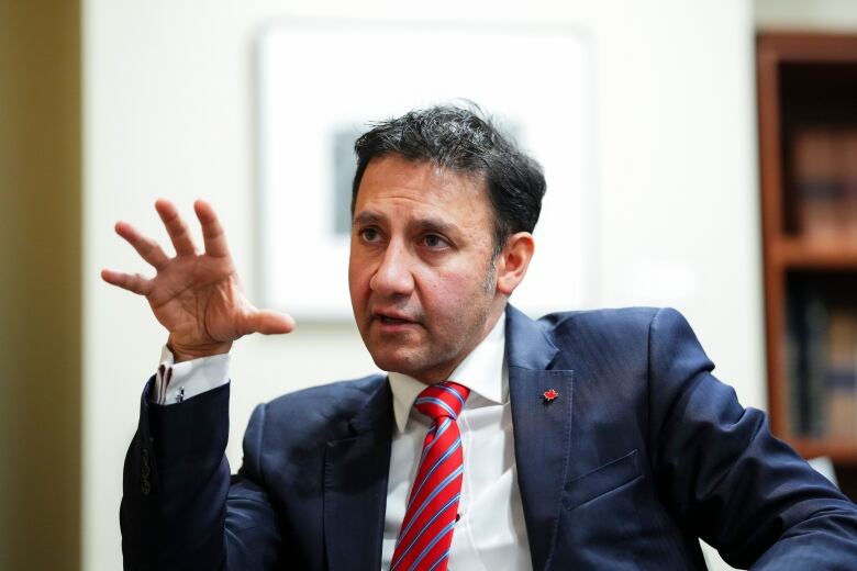 A man in a black tie and red suit gestures with his hand as he speaks.