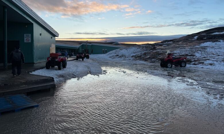 A large puddle is surrounded by ATVs on small amounts of snow and a mountain that has only patches of snow and bare ground. 