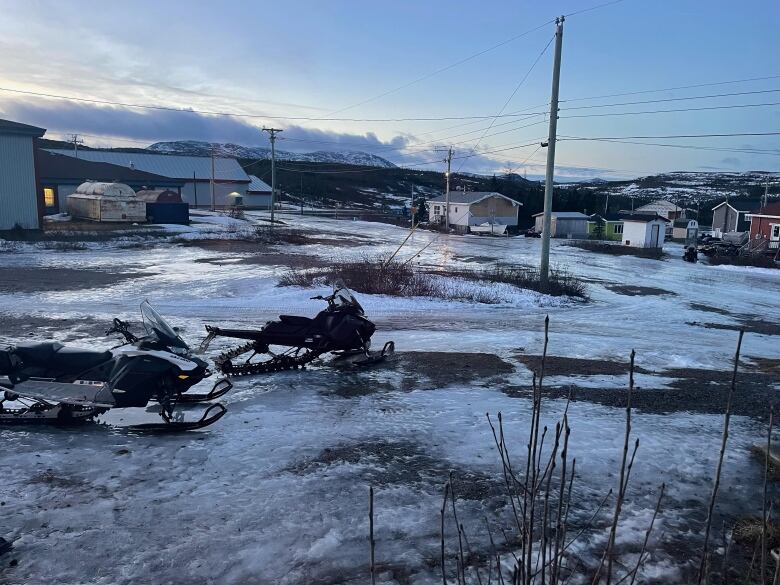 Two skidoos sit on ice and dirt with houses in the background. 