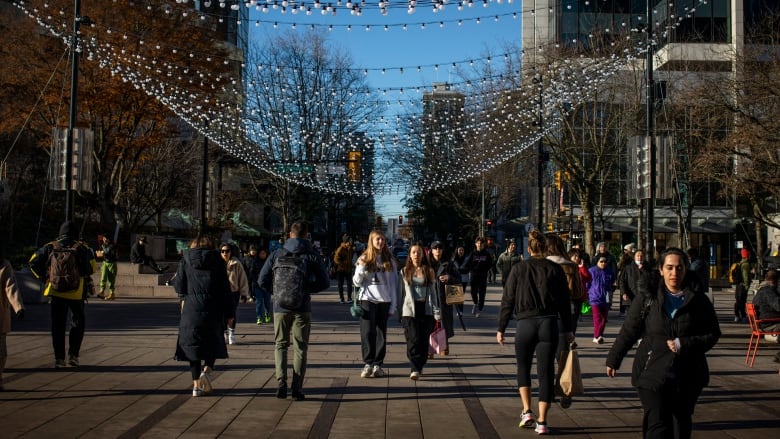 Dozens of people walking outside on a sunny fall day, including some who're passing under a canopy of Christmas lights.