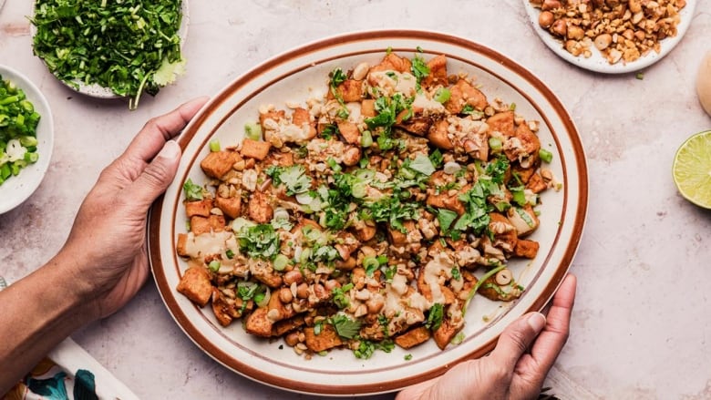 A roasted sweet potato salad with tahini dressing and topped with herbs on a serving platter held by two hands. 