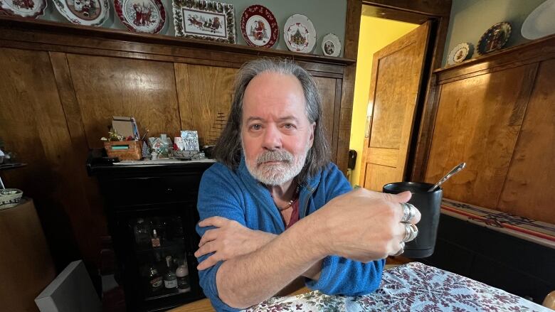 A man in a blue sweater holds a cup of coffee and stares at the camera while sitting at a table.