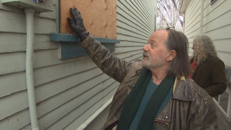 A man in a jacket holds his hand up to a boarded up window.