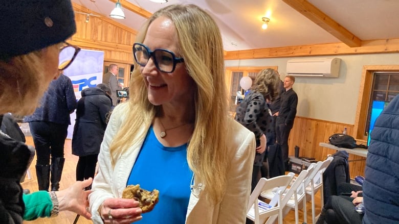 A smiling blond woman wearing a blue blouse and white blazer. She is holding a cookie in her hand.