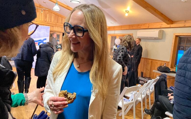 A smiling blond woman wearing a blue blouse and white blazer. She is holding a cookie in her hand.