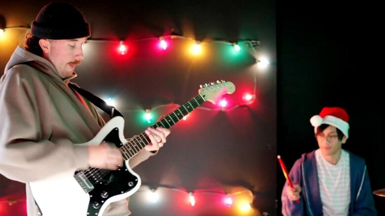 Man with a moustache wears a black beanie hat and plays a black and white electric guitar on the left side. There are two multi-coloured Christmas lights strung horizontally on a wall behind. A drummer wearing a red and white Santa hat plays on the right side. He is wearing a blue hoodie opened up with a white T-shirt with light grey horizontal stripes. 