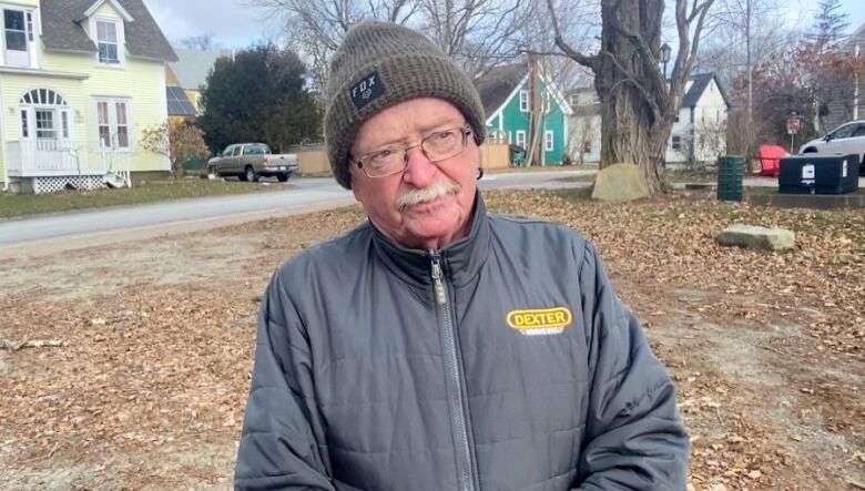 A white man with a moustache and glasses wears a black toque and black jacket, standing outside with colourful homes behind him