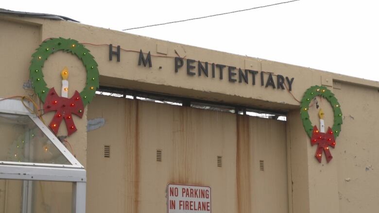 Two wooden Christmas wreath decorations bookend a metal sign on a yellow concrete building. The sign reads: H. M. Penitentiary.