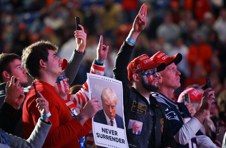 People in a crowd, some in red caps, hold up their phones and a sign that reads, 