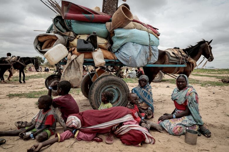 Several dark-complected people, including children, sit on the dirt in front of a horse that is carrying belongings.