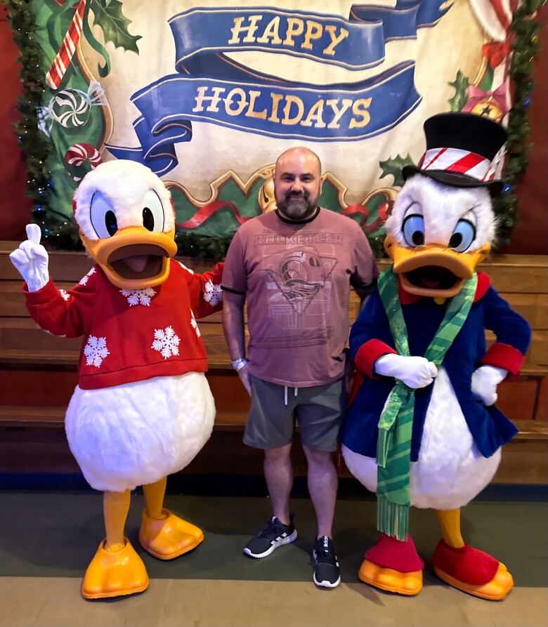 An adult man in a tshirt and shorts stands next to people dressed as Donald Duck and Scrooge McDuck.