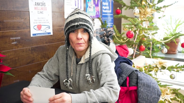 An older woman wearing a grey hoodie and a black woollen hat sits at a table.