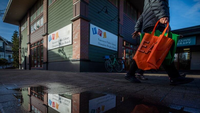 The outside of a wooden green building with a Vancouver Public Library sign. There are puddles on the ground and the building is reflected in them. A lone person walks past swinging a red tote bag and we can only see their black shoes and pants.