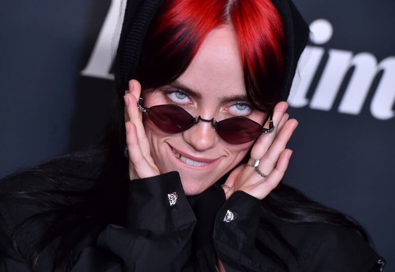 A woman with red hair in a black dress and sunglasses smiles for photographers.