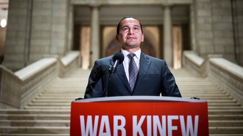 A man in dark hair and a suit stands at a podium, at the base of a grand staircase.