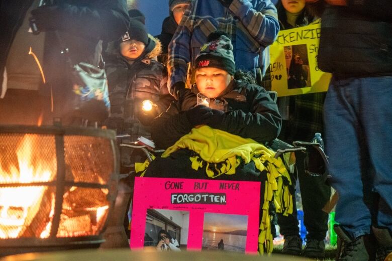 A young person covered in blankets is seen holding a candle by a fire.