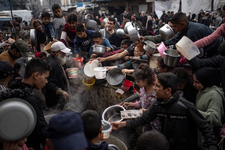 A crowd of people hold out bowls as they surround a person distributing food.