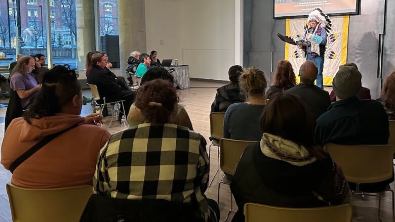 a few dozen people sit in chairs listening to a man wearing an Indigenous headdress speak at a podium