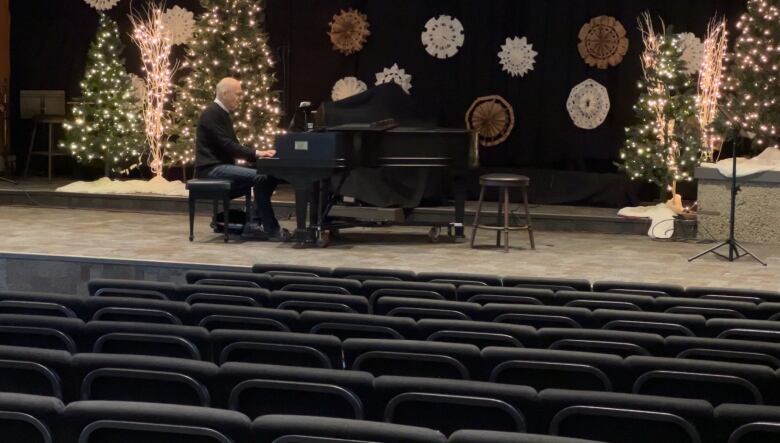 A man in his 60s sits at a grand piano on a stage in a large and empty church in Penticton, practicing the piano.