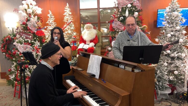 A musical trio performs a Christmas concert at a hotel lobby surrounded by festive decorations. A middle aged woman is standing and singing at a microphone beside a man in his 60s playing the piano and a middle aged man nearby playing the bass.