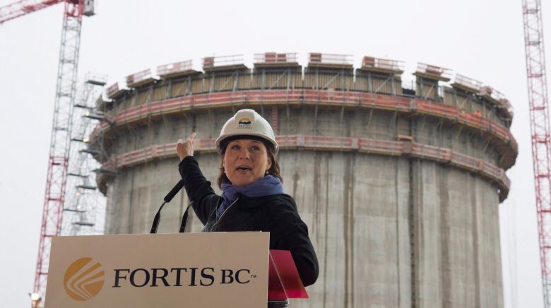 A woman wearing a hard hat, standing at a podium marked 'FortisBC', points to a large under-construction tower behind her.