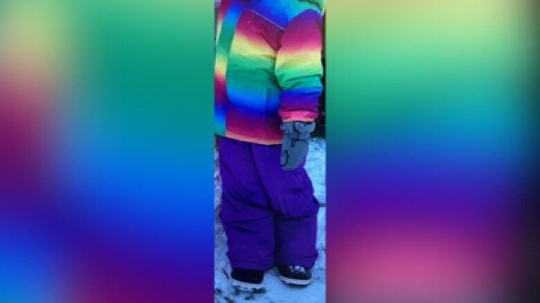 Photo of a girl wearing a rainbow-gradient winter jacket and blue snow pants. Her head is cropped. 