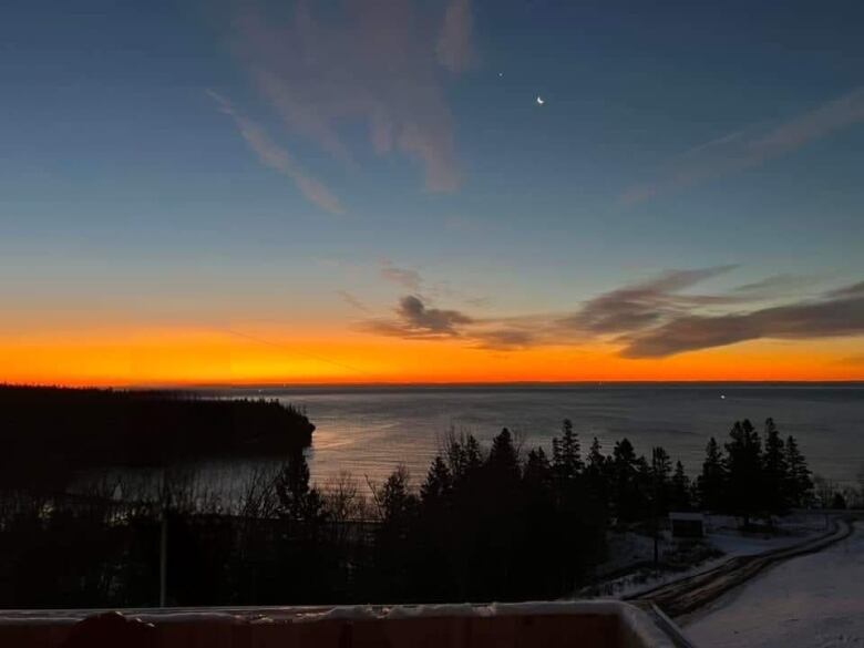 An orange sunrise over a large expanse of water. A crescent moon is visible high up in the sky.