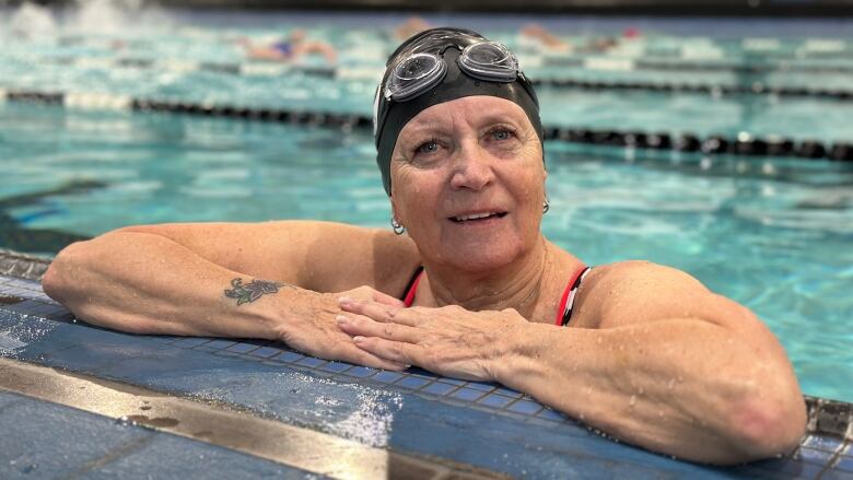 A woman stands inside a swimming pool wearing a swim cap with goggles above her eyes. Her arms are folded on the side of the pool.