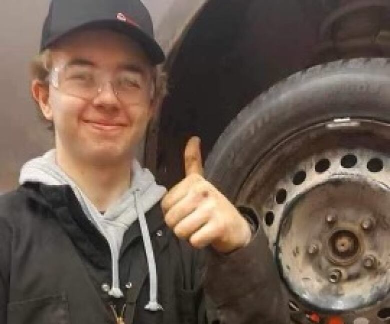 A young man in overalls and a baseball cap gives a thumbs up in front of a car in a mechanic shop.