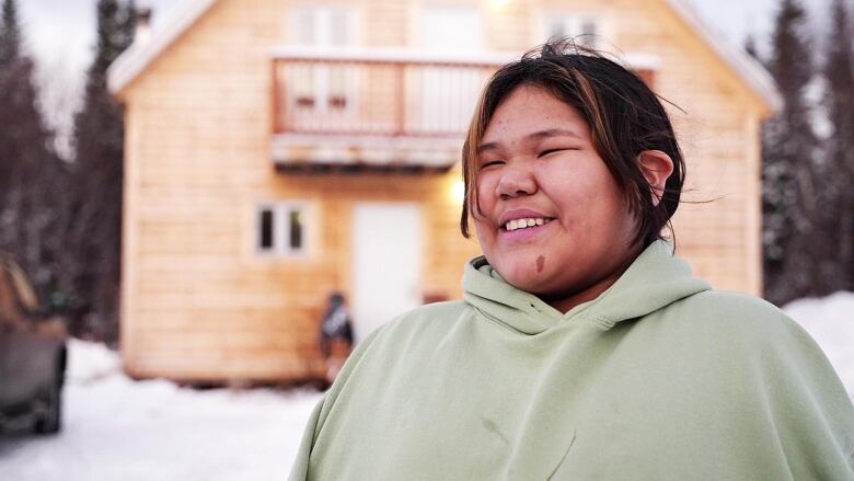 A young girl in a green sweater stands in front of a wooden, two-storey cabin. 