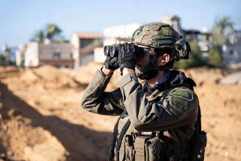 A soldier in full gear including a helmet looks through binoculars amid a brown landscape. Buildings are visible out of focus in the background.