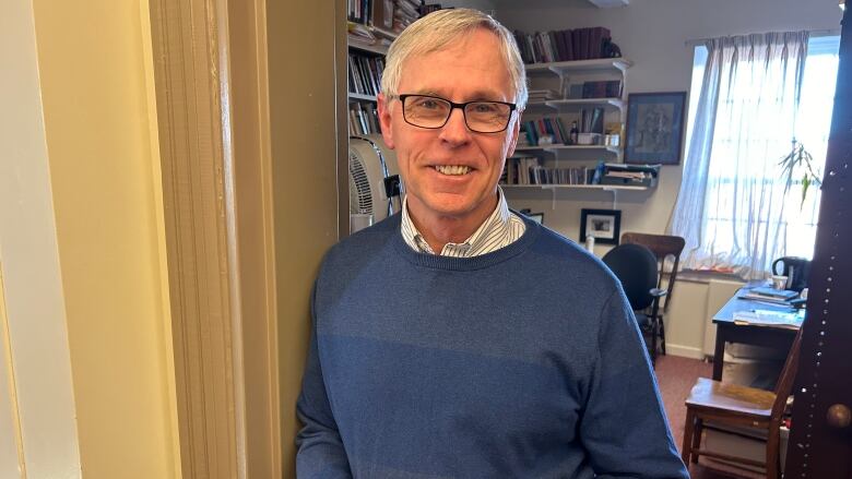 A man in a blue sweater over a white collared shirt stands in the doorway of an office.