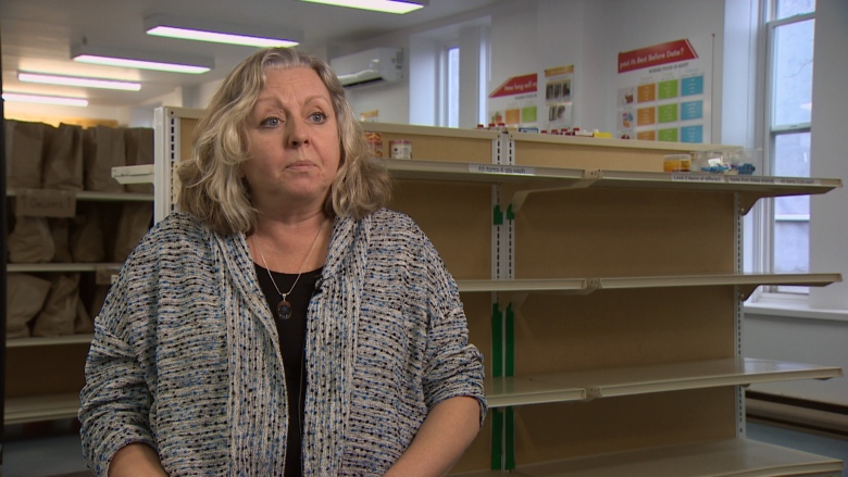 A woman with light, curly hair wears a grey cardigan. She is sitting in front of empty shelves. 